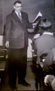 A photo of Alva Cavett standing in front of a classroom of students, reading from a book in his hand